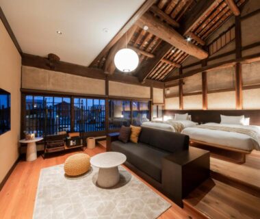 Spacious loft-style room with exposed beams, twin beds on raised platform, seating area and large windows at Tomonoura Shiomachi Hotel