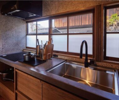 Compact kitchenette with stainless sink, stovetop, wooden cabinets and frosted window at Tomonoura Shiomachi Hotel, Hiroshima