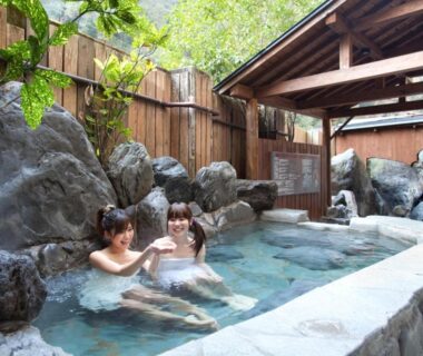 Two guests soaking in a rock-lined onsen bath with wooden fence and shelter at Tsuetate Onsen Hotel Hizenya, Kumamoto