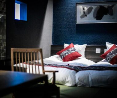 Low twin beds with cushions, wooden chair and wall art in a dimly lit Tsukasa Ryokan room, Saga, Kyūshū