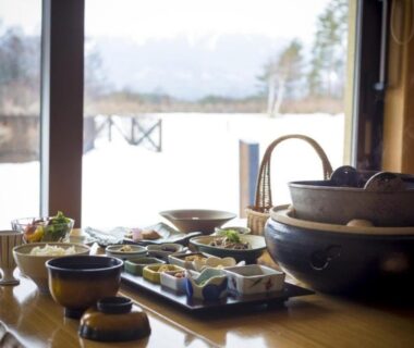 Low wooden table set with assorted Japanese dishes and ceramic pots, snowy landscape visible through window