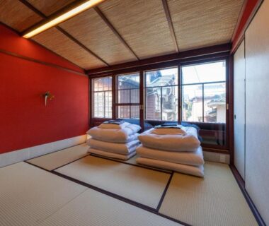 Tatami room with stacked futons, shoji windows and red accent wall at UTAIMACHI in Chūbu, Ishikawa, Japan