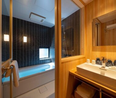 Wooden vanity and sink, glass-partitioned bathtub and tiled wall in UTAIMACHI ryokan bathroom, Ishikawa, Chūbu