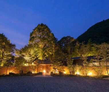Dusk view of illuminated stone entrance and lanterns with trees and mountain backdrop at Ufufu Shizuoka