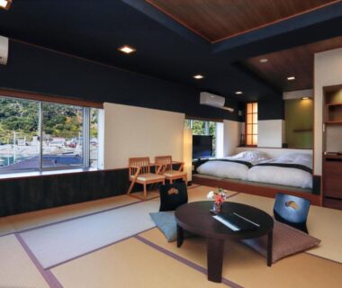 Tatami room with low table, floor chairs, two beds and large windows showing street and greenery at Uminoniwa Chiba