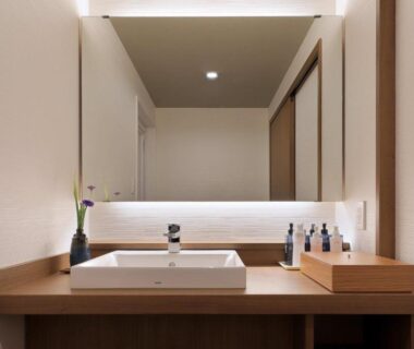 Wooden vanity with rectangular sink, illuminated mirror, toiletries and small vase on the counter