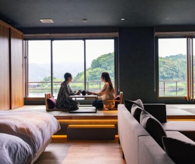 Two guests in yukata at low table in a guestroom with beds, sofa and windows overlooking green hills