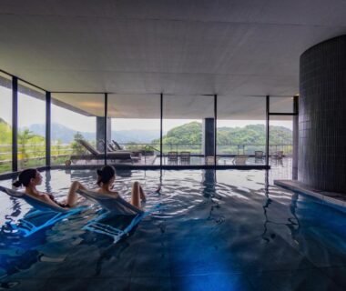 Guests relaxing in indoor onsen pool with floor-to-ceiling windows and mountain view at Urari Takeo Garden Terrace