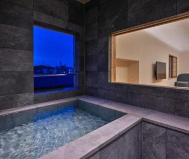Stone-tiled indoor onsen bath with large window and interior room visible through glass partition