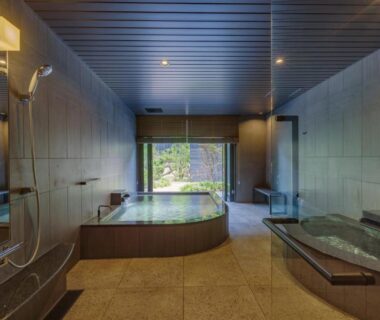 Tiled indoor onsen with wooden soaking tub, bench and window opening to a framed garden view