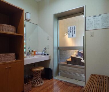 Changing area with vanity sink, wicker baskets, bench and wooden bathing tub behind sliding door at Uruoikan Nagano