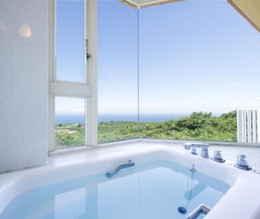 Indoor soaking tub beside panoramic window overlooking green hillside and distant sea at Village Izukogen Shizuoka