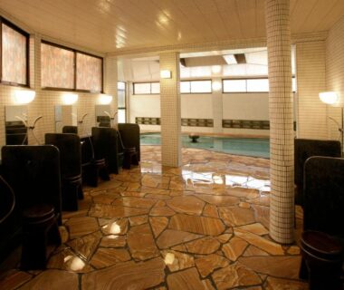 Indoor communal onsen with tiled stone floor, seated washing stations and central bathing pool at a Nagano ryokan