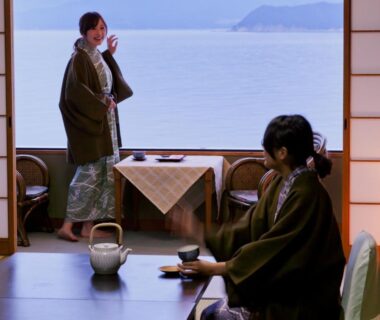 Two guests in yukata in tatami room with low table, tea set and sea visible through shoji screens