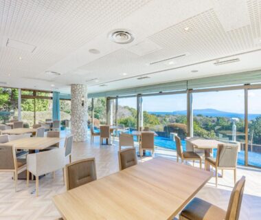 Bright dining area with wooden tables and chairs, floor-to-ceiling windows overlooking green hillside and distant horizon
