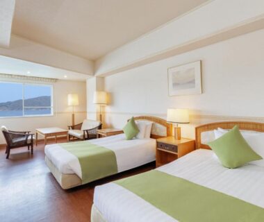 Twin beds with green cushions, bedside lamps, seating area and window overlooking coastal waters in Wan Wan Paradise Toba Mie