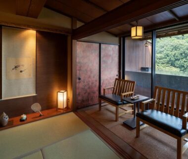 Tatami room with tokonoma alcove, low table, wooden chairs and balcony overlooking green hillside in Shizuoka