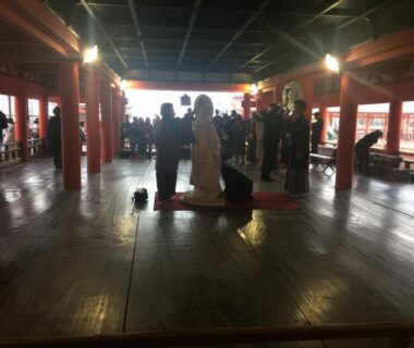 Polished wooden hall with pillars and kneeling visitors at Wholeearth Ryokan Hiroshima in Chūgoku, Hiroshima, Japan