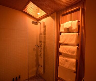 Warm-lit shower area with glass door and stacked towels on wooden rack at Yado Kiramachi, Kyoto