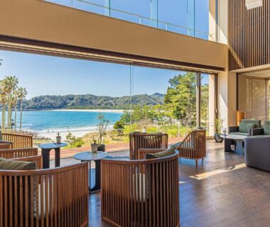 Open lounge with wooden seating and floor-to-ceiling windows overlooking sandy beach and coastal waters in Shizuoka