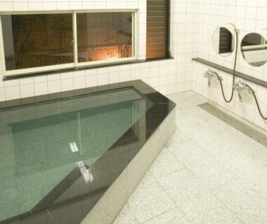 Tiled indoor bathing room with rectangular soaking tub, wall-mounted shower stations, mirrors and bench at Yadoya Nishijinso, Kyoto