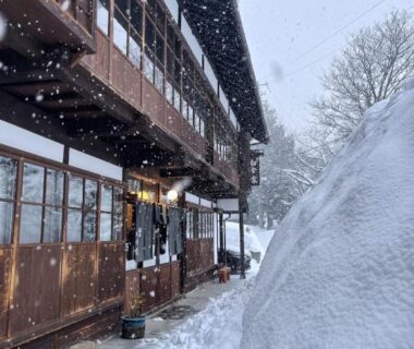 Wooden multi-story ryokan exterior under heavy snowfall with large snowbank at entrance in Nagano