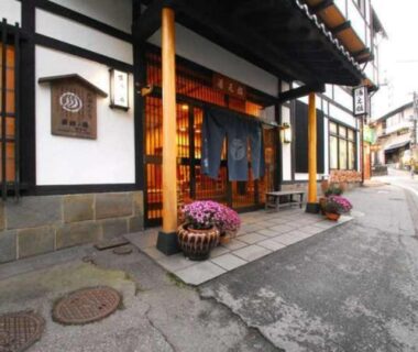 Traditional ryokan entrance with noren curtains, wooden facade and potted flowers along a narrow street in Gunma