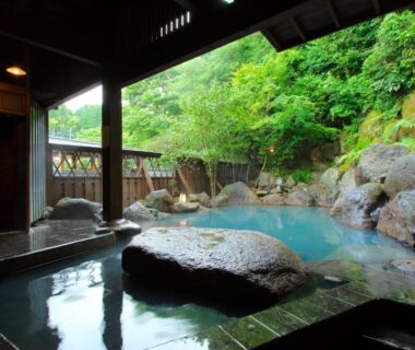 Rock-lined outdoor onsen with covered wooden edge and lush green hillside at Yamabiko Ryokan Kumamoto