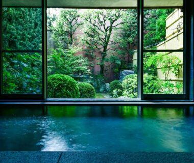 Indoor onsen bath with low ledge overlooking lush Japanese garden through large windows
