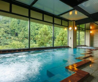 Indoor hot spring pool with large windows overlooking forest, tiled steps and wooden accents at Yamanaka Onsen Hanatsubaki