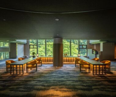 Spacious ryokan lobby with low wooden tables, cushioned chairs, central pillar and large windows framing verdant trees