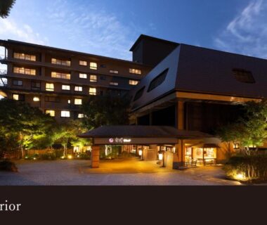 Dusk view of lit ryokan entrance and multi-storey exterior with landscaped trees at Yamanaka Onsen, Ishikawa