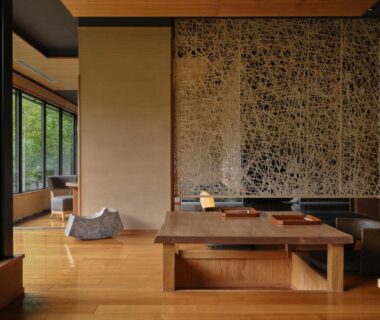 Interior seating with low wooden table, cushions, woven wall panel and garden-facing windows at Yamanakaonsen Hanamurasaki, Ishikawa