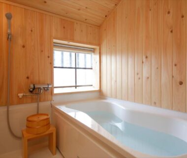 Wood-paneled bathroom with filled bathtub, shower head, wooden stool and small window at a Hyōgo ryokan.