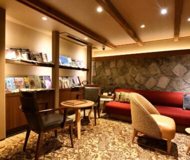 Warm ryokan lounge with bookshelves, seating area, stone accent wall and wooden beams at Yasaka Yutone Kyokoyado in Kyoto
