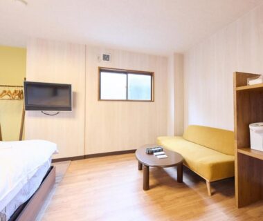 Bright room with Western bed, yellow sofa, low table, wall-mounted TV and wooden flooring at Yodoya in Hyōgo