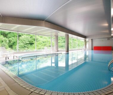 Tiled indoor bathing pool with large windows overlooking lush greenery and poolside deck chairs