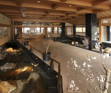 Spacious wooden-beamed ryokan lobby with stone water feature and flowering branches at Yoshikawaya Fukushima