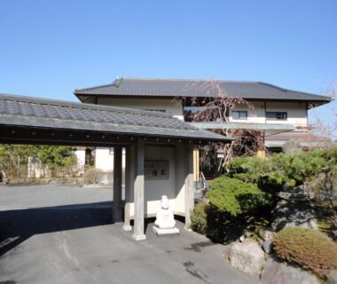 Covered entrance gate and driveway leading to Yoshimatsu ryokan with tiled roofs and manicured shrubs in Kanagawa