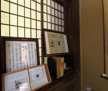 Shoji window with framed documents and books on wooden ledge beside hanging scroll in ryokan room