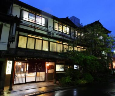 Lit wooden ryokan façade with shoji windows and entrance on wet street at Yudanaka Yumoto, Nagano