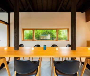 Long wooden communal table with black chairs and low windows framing lush green garden at a ryokan dining space