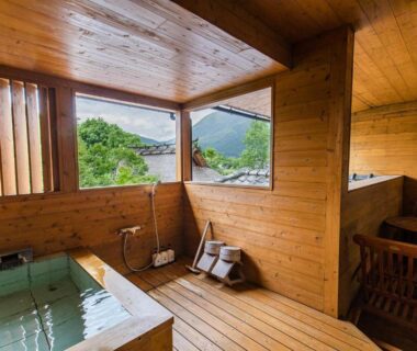 Indoor wooden bath with windows framing green hills, wooden floor and seating at Yufuin ryokan, Ōita