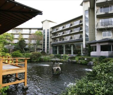 Pond and wooden terrace with multi-story ryokan building in courtyard at Yufuin Kotobuki Hananosho, Ōita
