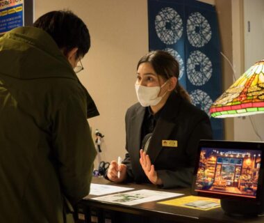 Guest speaking with masked receptionist at Yufuin Lamp no Yado reception, Ōita, Kyūshū
