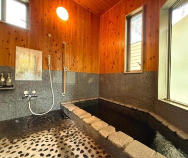Indoor stone onsen bath with wooden panel walls, shower fixtures and pebble floor at Yufuin Onsen Yaeichi, Oita