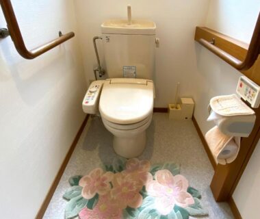 Compact toilet with washlet controls, wooden handrails and floral floor mat at Yufuin Onsen Hasuwa Inn, Ōita