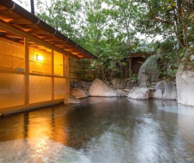 Stone-lined outdoor onsen with wooden veranda and ambient lantern lighting surrounded by trees at a Yufuin ryokan