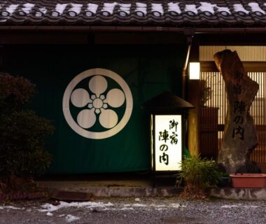 Traditional ryokan entrance with green noren emblem, illuminated signboard and stone name pillar in Ōita, Kyūshū
