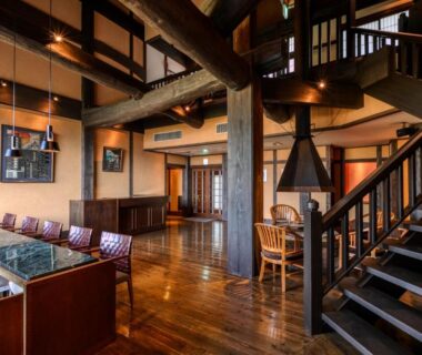 Spacious wooden lobby with exposed beams, seating area, staircase and suspended fireplace at Yufuin Onsen Toshoan, Ōita